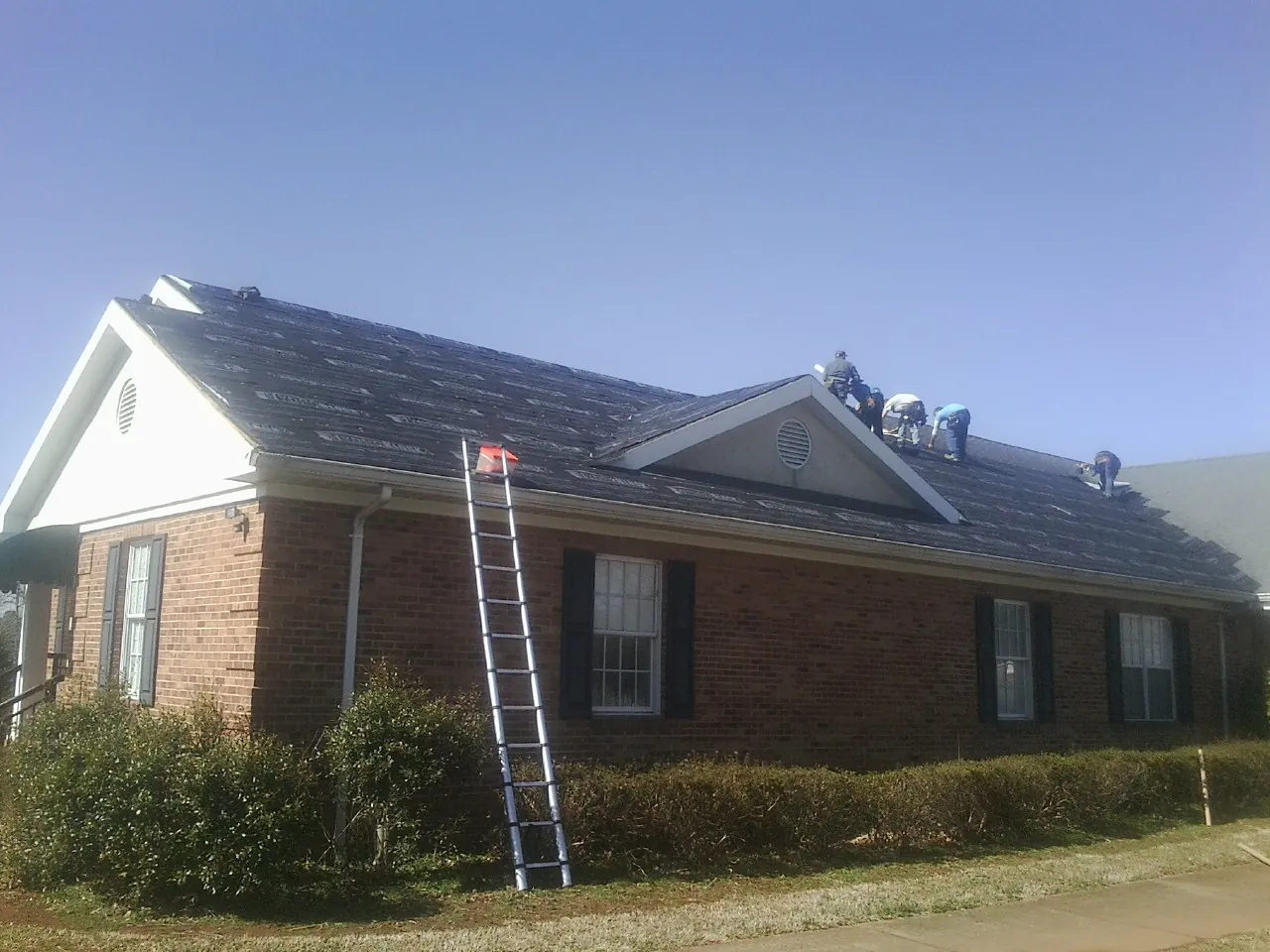 Crew tearing shingles off a brick ranch roof with a ladder set — Emergency Roof Repair work by our crew in Rumney