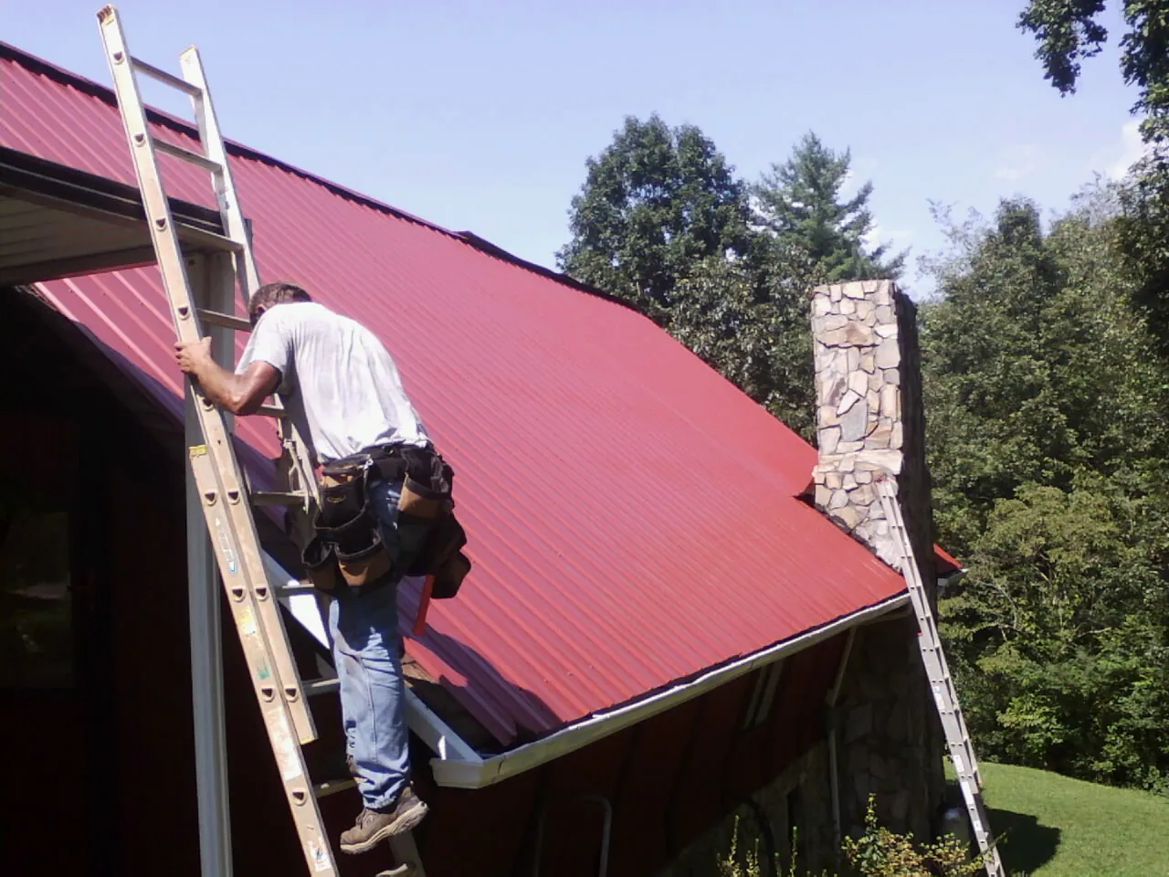 Roofer on a ladder beside a red metal roof and stone chimney — Soffit & Fascia Repair by Roofing Brothers in Rumney